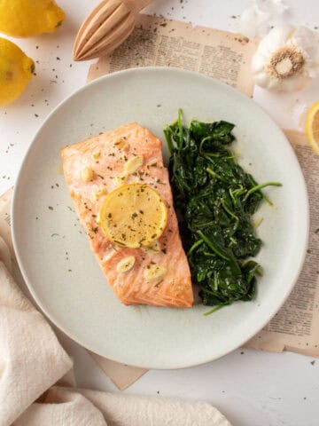 Two filets of salmon topped with a lemon, butter, garlic sauce on a plate with sauteed spinach surrounded by lemons and a napkin.