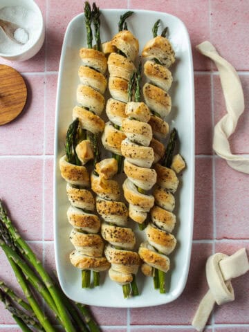 Asparagus wrapped puff pastry rolls on a white platter with a pink tiled background surrounded by asparagus and pastry dough.