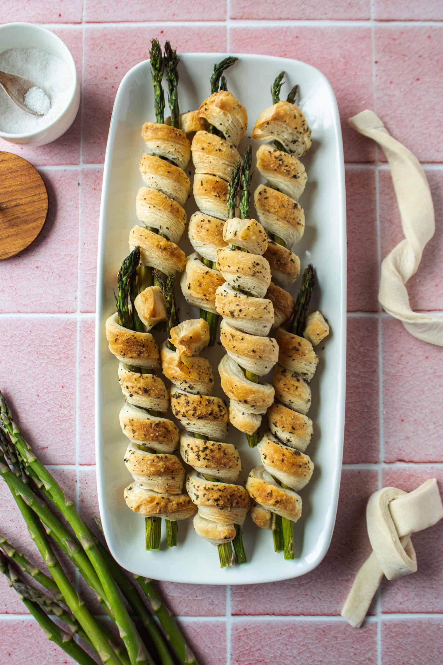 Asparagus wrapped puff pastry rolls on a white platter with a pink tiled background surrounded by asparagus and pastry dough.