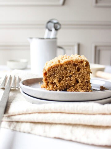 A slice of a banana crumb cake topped with a streusel topping on a plate next to a fork with a napkin under it.