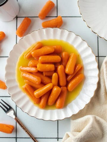 Pineapple candied baby carrots on a small scalloped white plate with an ivory napkin next to it with scattered carrots surrounding the plate.