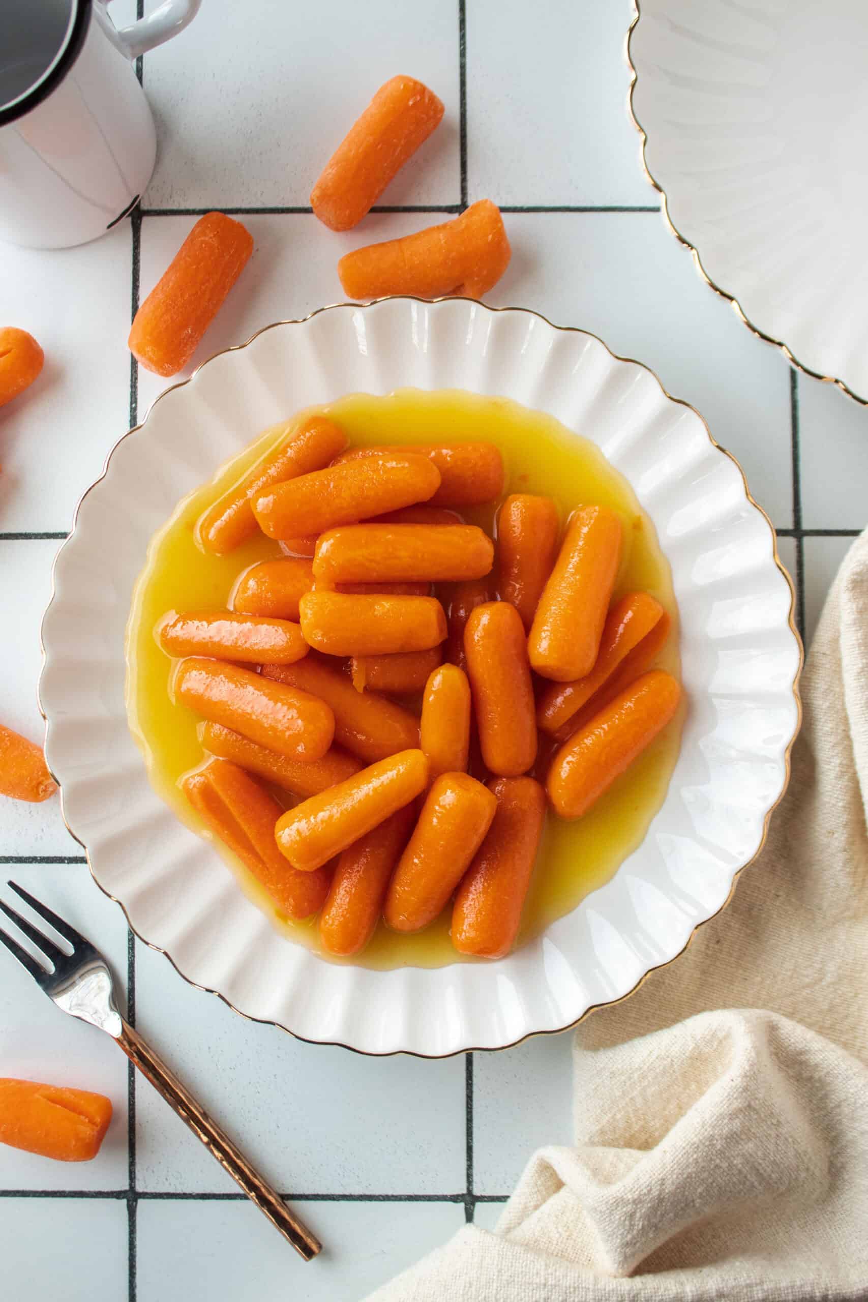Pineapple candied baby carrots on a small scalloped white plate with an ivory napkin next to it with scattered carrots surrounding the plate.