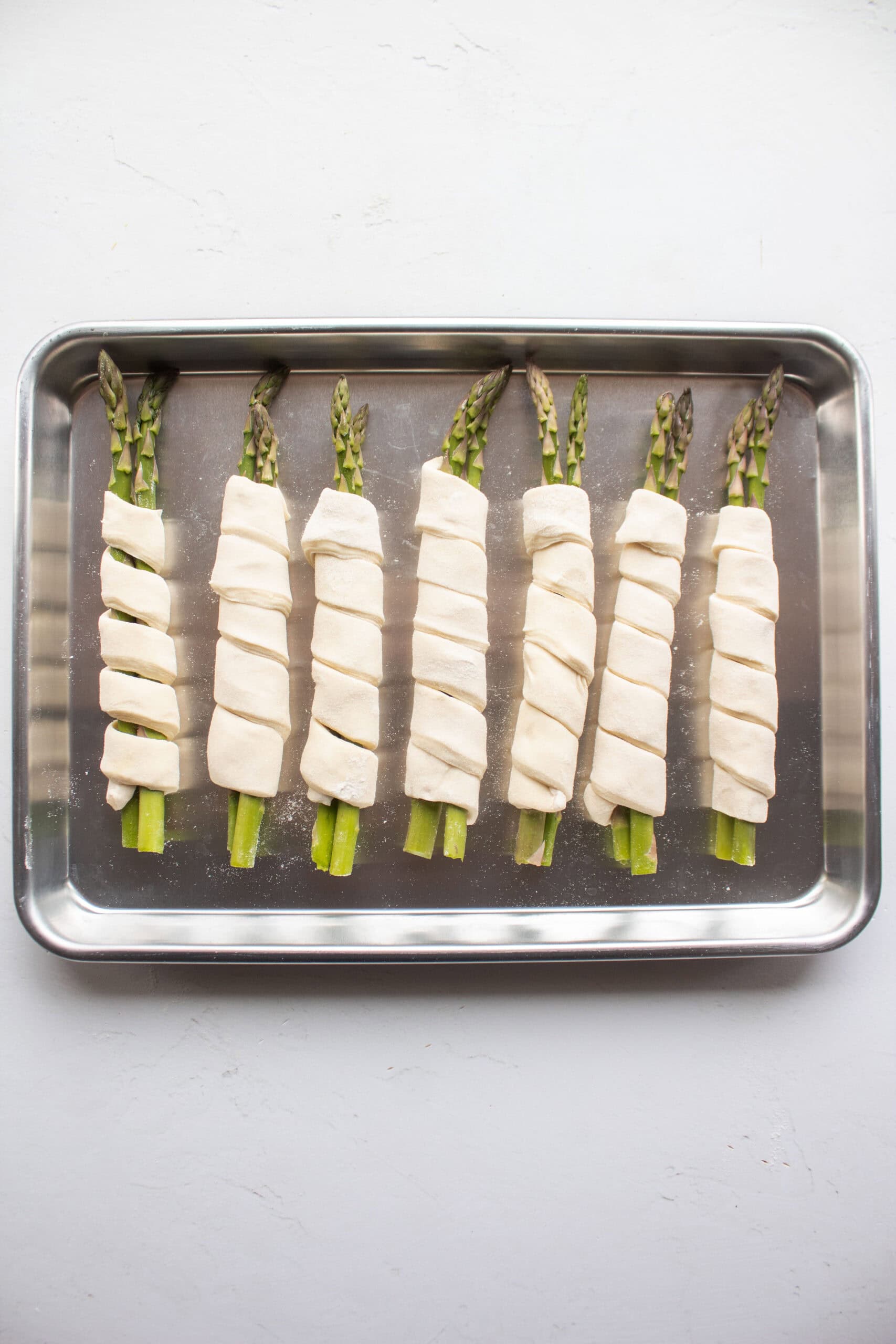 Seven pieces of asparagus wrapped in puff pastry on a baking sheet.