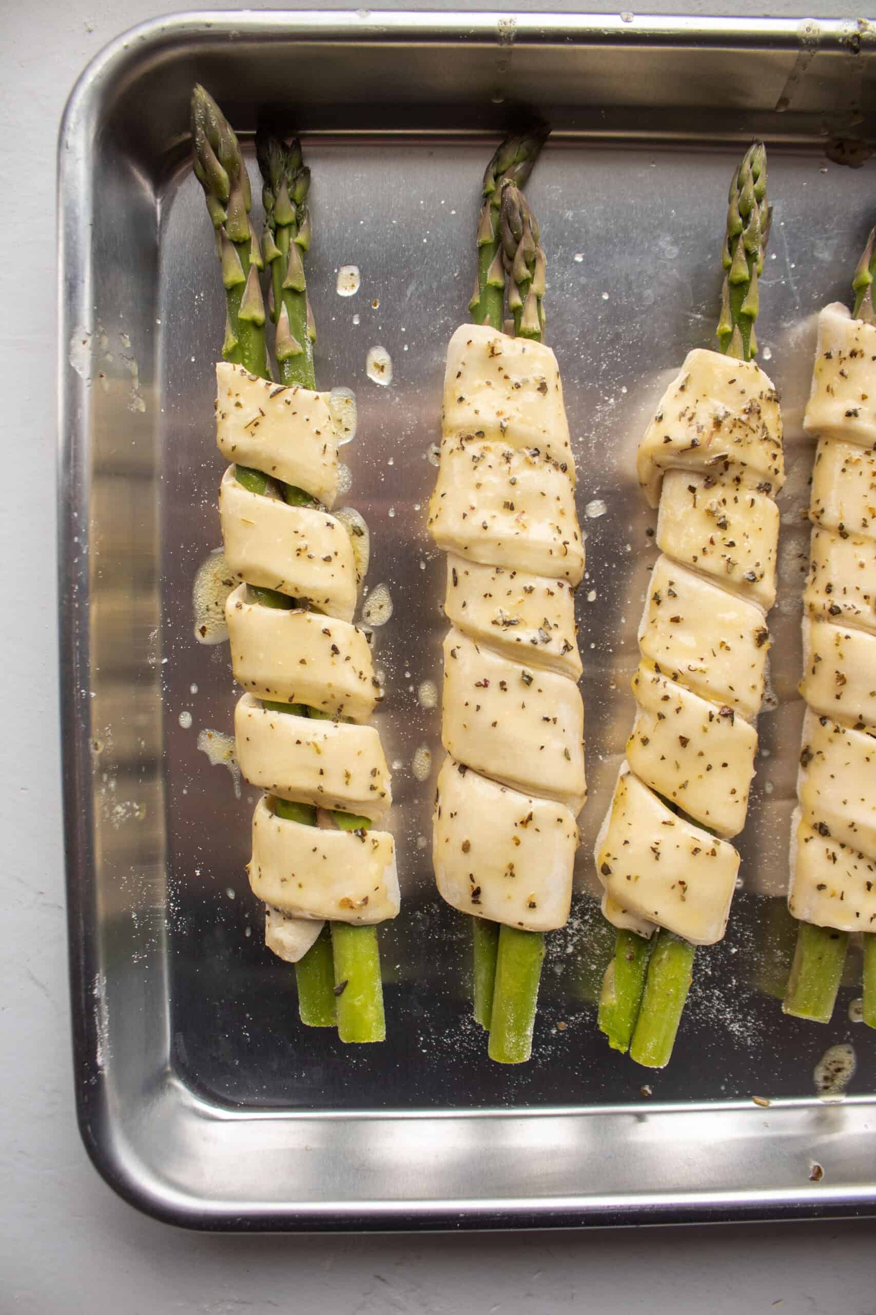 Seven pieces of asparagus wrapped in puff pastry on a baking sheet topped with melted butter and dried basil.