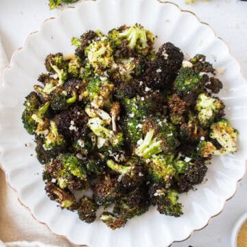 Crispy air fryer broccoli with parmesan cheese on a white scalloped plate surrounded by loose broccoli florets and a napkin.
