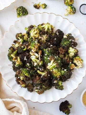 Crispy air fryer broccoli with parmesan cheese on a white scalloped plate surrounded by loose broccoli florets and a napkin.