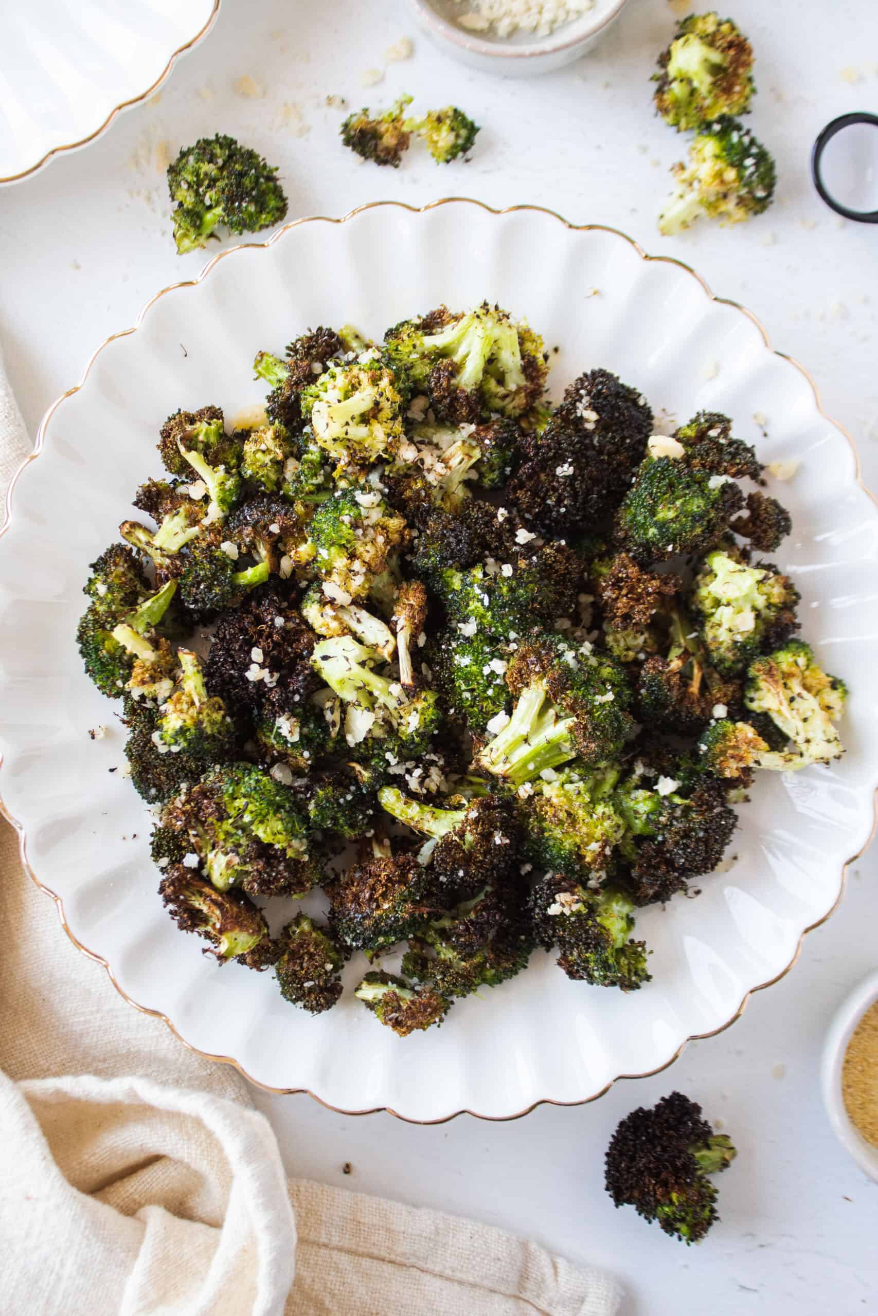 Crispy air fryer broccoli with parmesan cheese on a white scalloped plate surrounded by loose broccoli florets and a napkin.