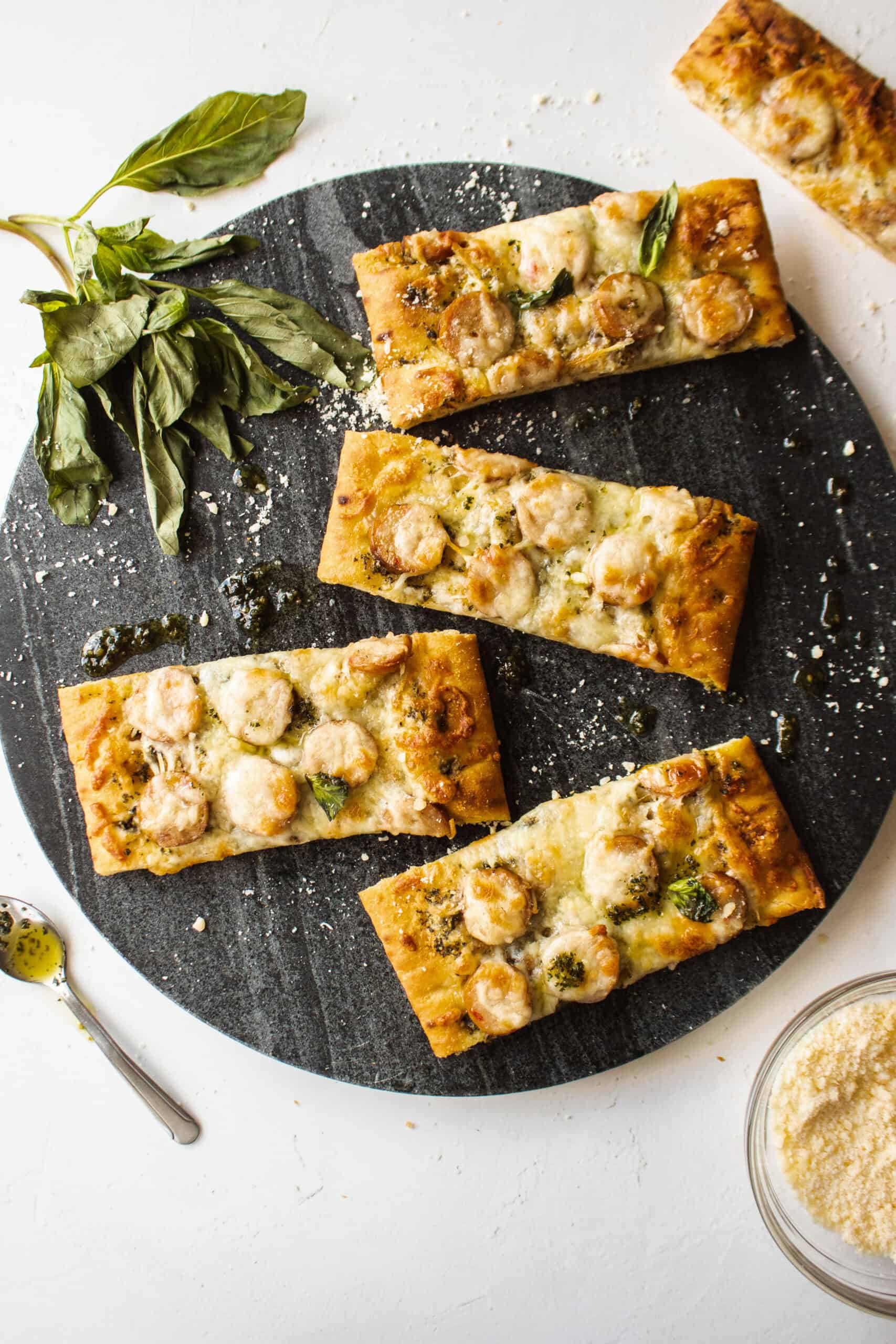 Four slices of a chicken sausage and pesto flatbread on a serving platter surrounded by basil and parmesan cheese.