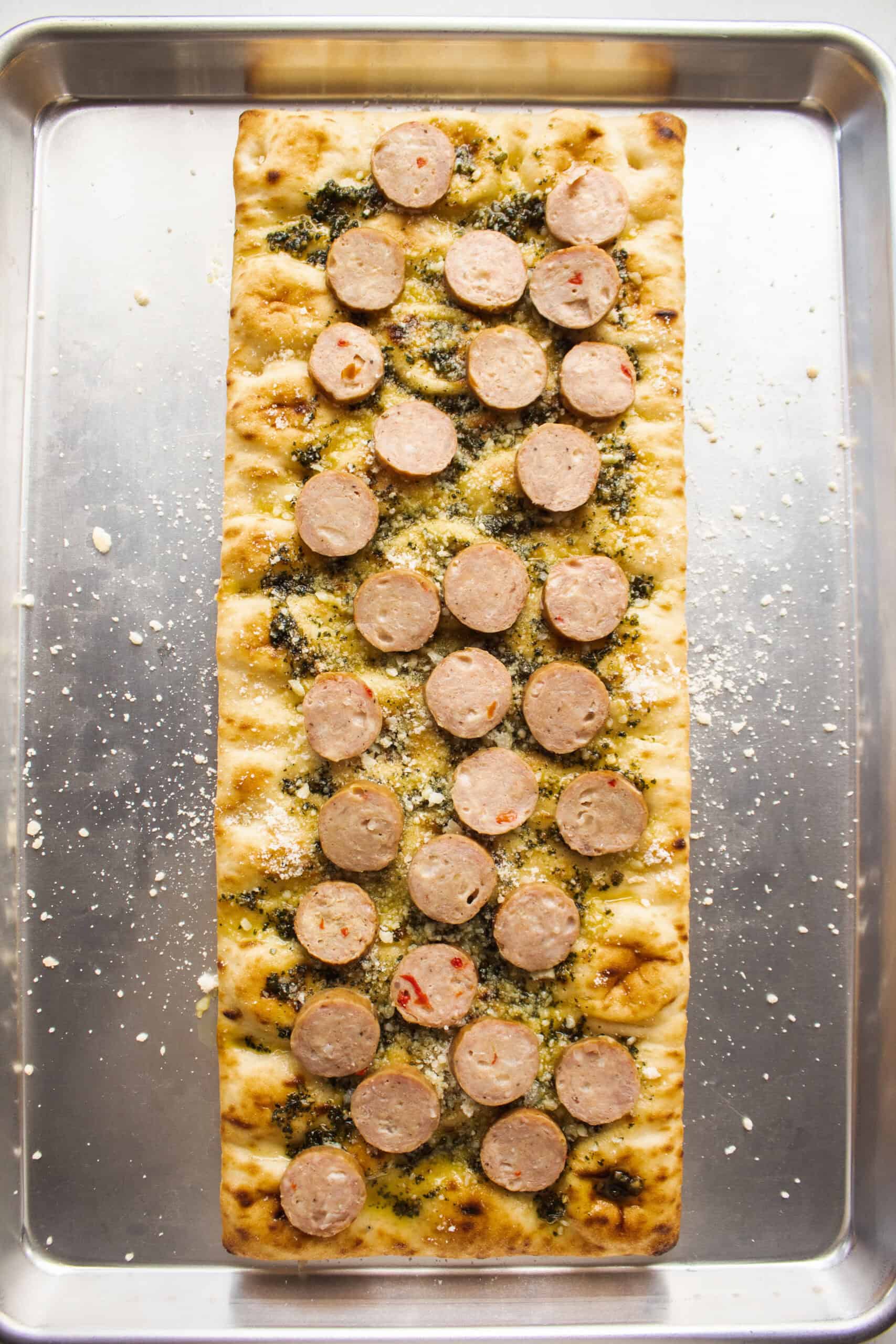 Pesto spread on a flatbread on a sheet pan topped with parmesan cheese and sliced chicken sausage.