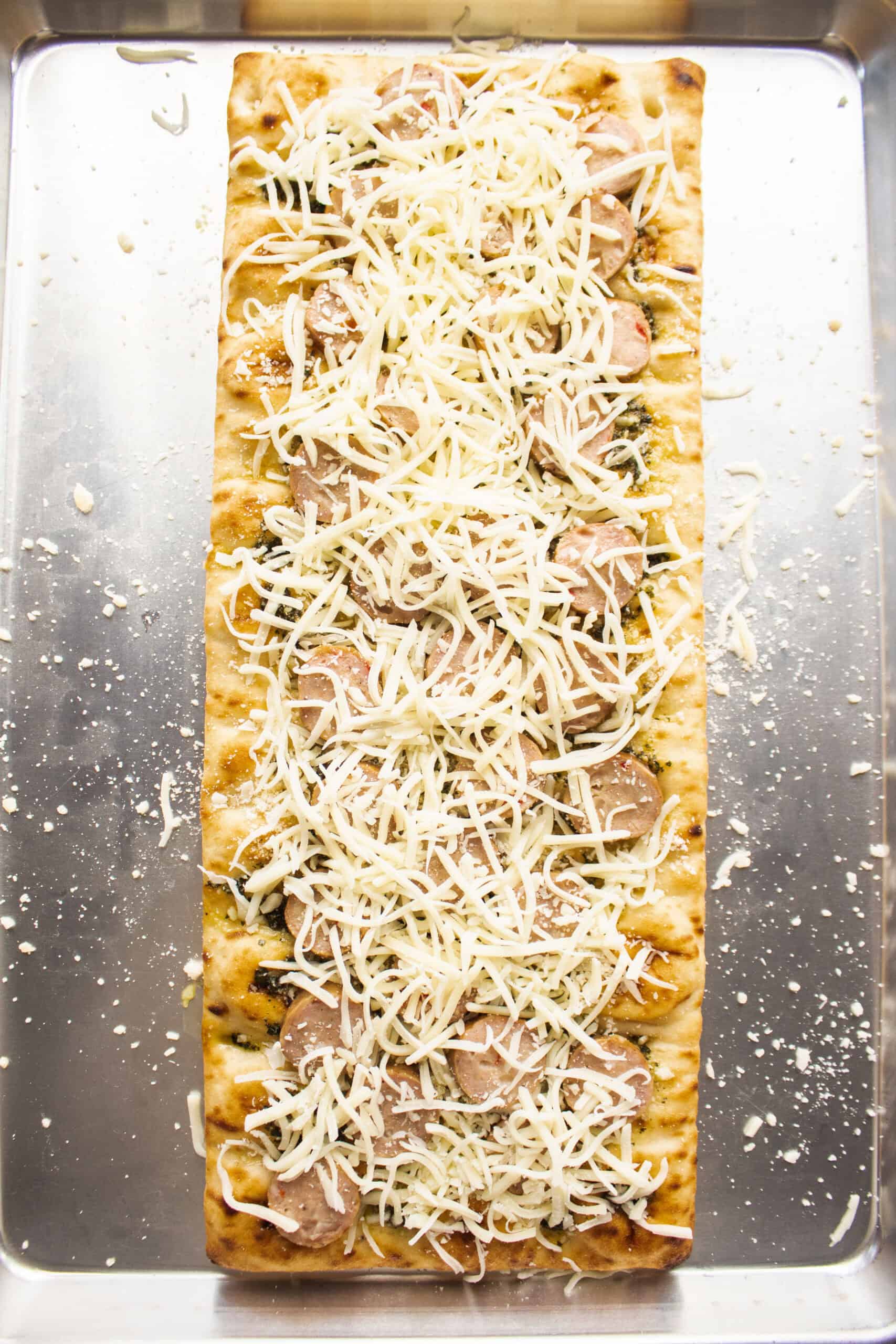 Pesto spread on a flatbread on a sheet pan topped with parmesan cheese, sliced chicken sausage, and mozzarella cheese.