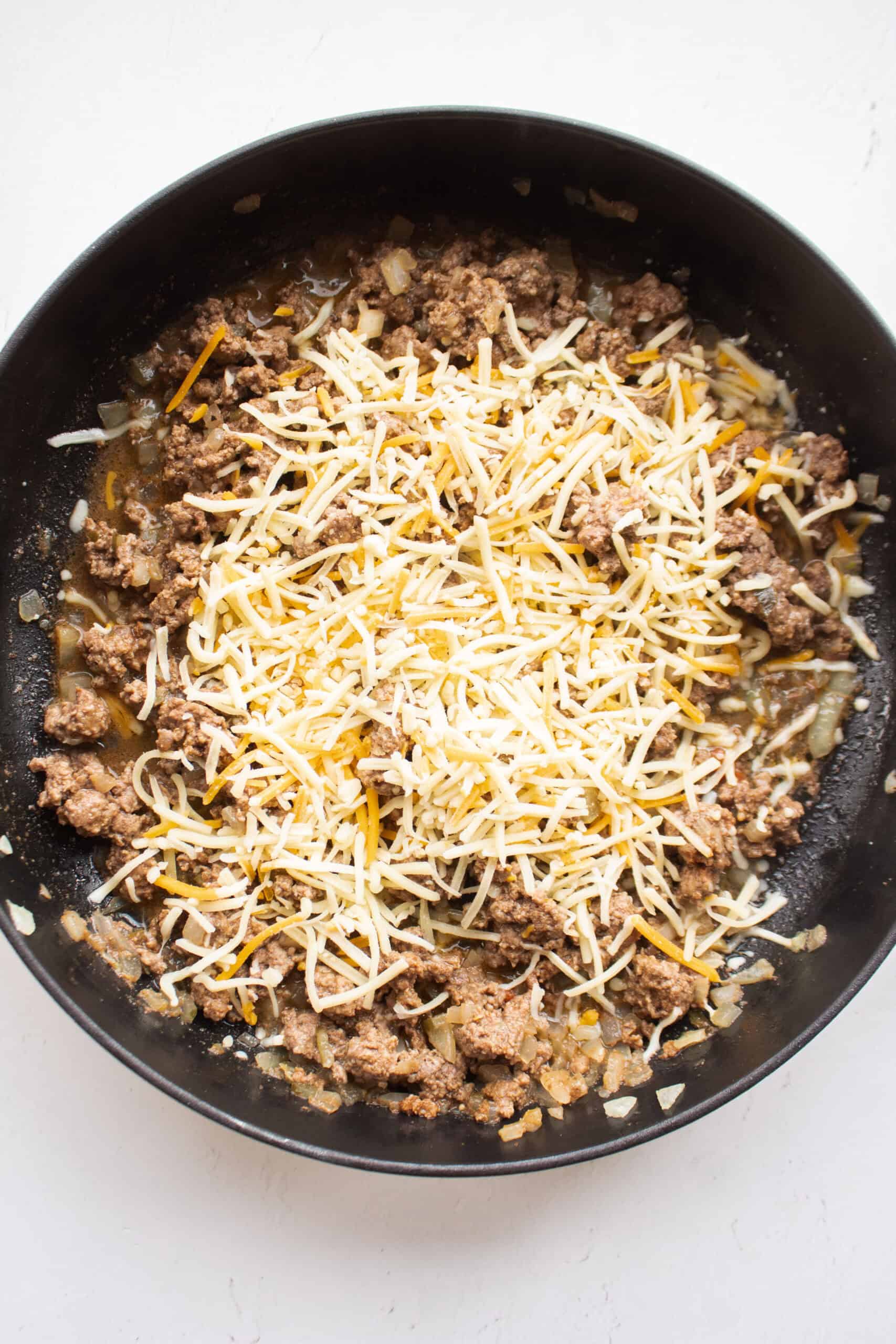 Shredded monterey jack cheese on top of ground beef seasoned with garlic, paprika, salt, and pepper combined with sauteed onions in a skillet.