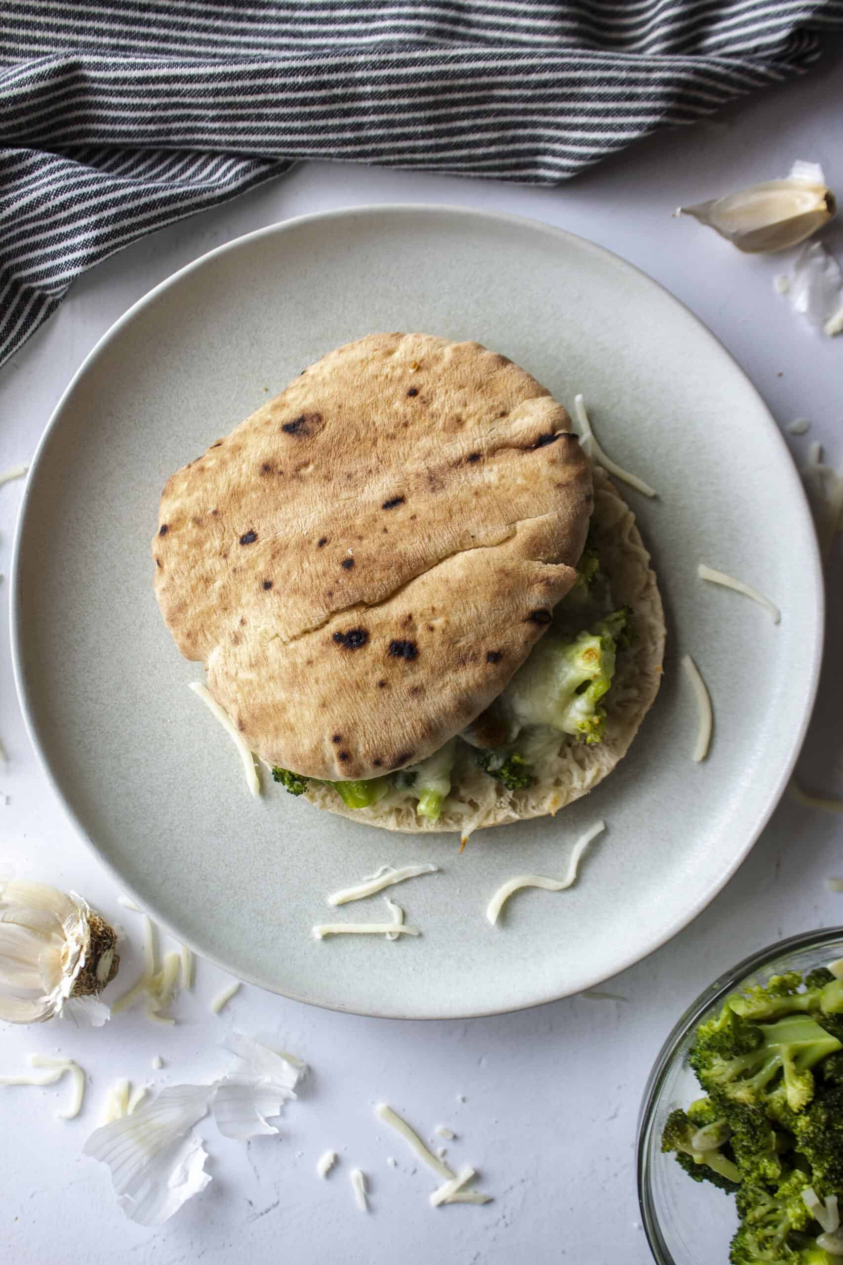 A broccoli pita melt sandwich on a gray blue plate surrounded by cheese, more broccoli, and a blue and white striped napkin.