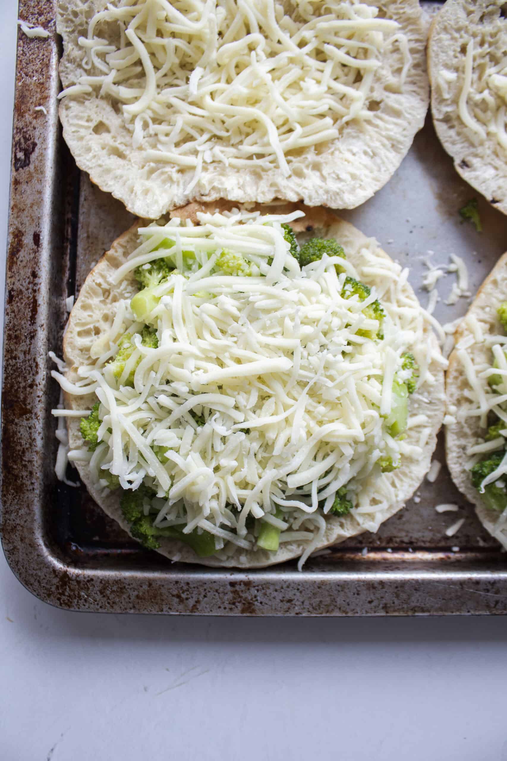 A pita bread sliced open on a small baking sheet with one side topped with cheese and the other with sautéed broccoli and more cheese.