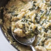 Close up of a spoon in a cast iron skillet holding a spoonful of creamy orzo with chicken and kale.