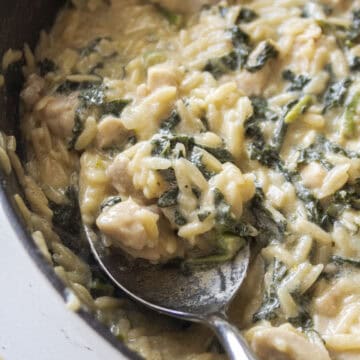 Close up of a spoon in a cast iron skillet holding a spoonful of creamy orzo with chicken and kale.