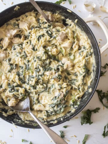 Creamy orzo with chicken and kale in a cast iron skillet with two serving spoons inserted on either side surrounded by shredded kale and garlic.