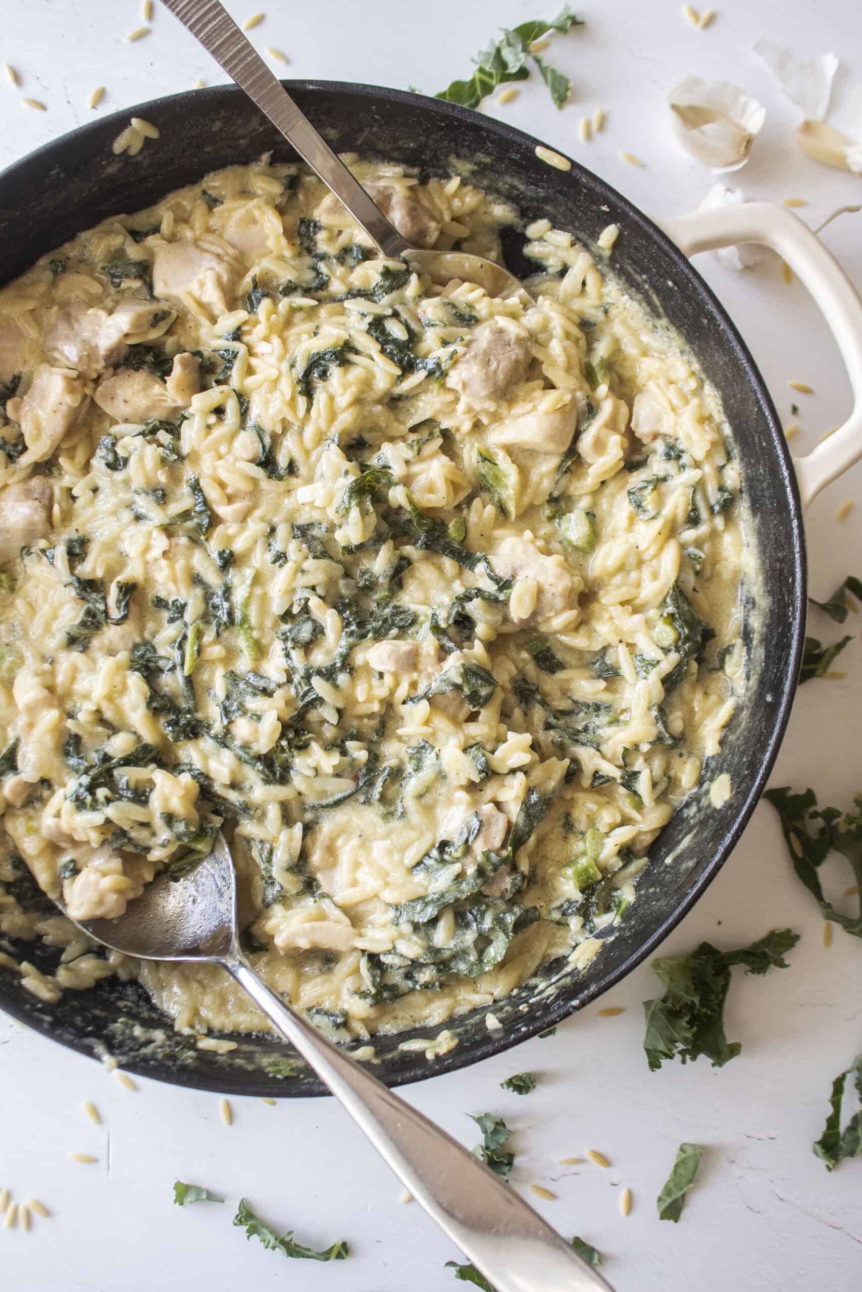 Creamy orzo with chicken and kale in a cast iron skillet with two serving spoons inserted on either side surrounded by shredded kale and garlic.
