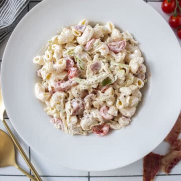 BLT pasta salad in a large bowl surrounded by cherry tomatoes, bacon, and serving spoons.