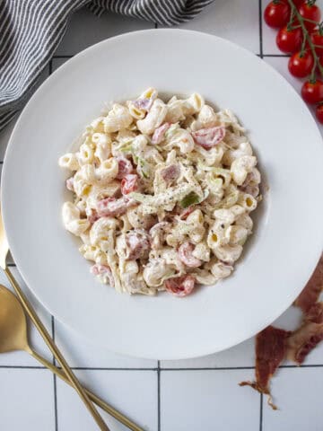 BLT pasta salad in a large bowl surrounded by cherry tomatoes, bacon, and serving spoons.
