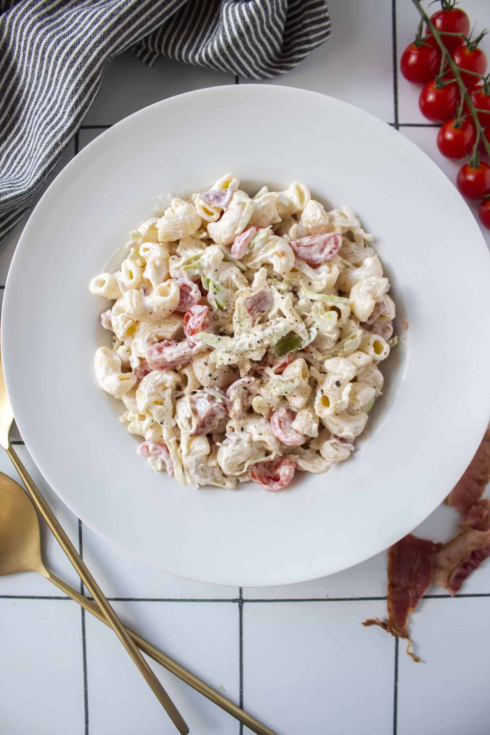 BLT pasta salad in a large bowl surrounded by cherry tomatoes, bacon, and serving spoons.