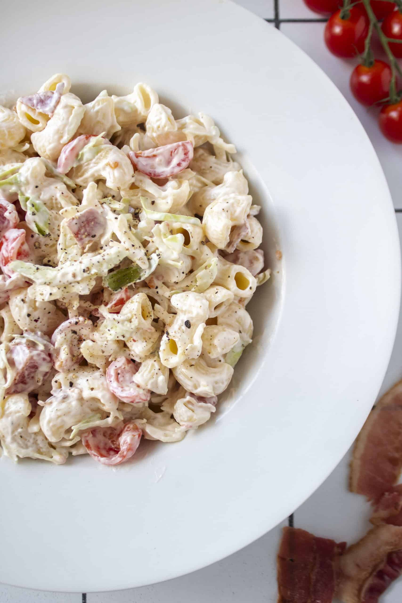 Close up of BLT pasta salad in a large bowl surrounded by cherry tomatoes and bacon.