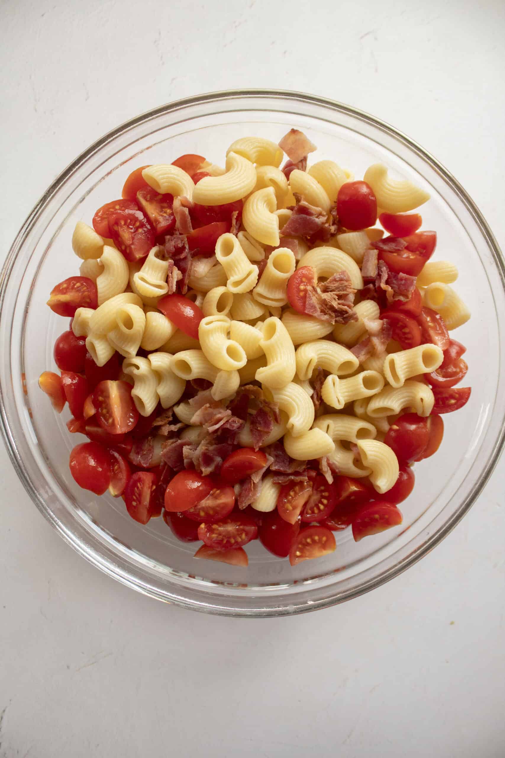 Cooked elbow macaroni in a large glass mixing bowl with quartered tomatoes and diced bacon.
