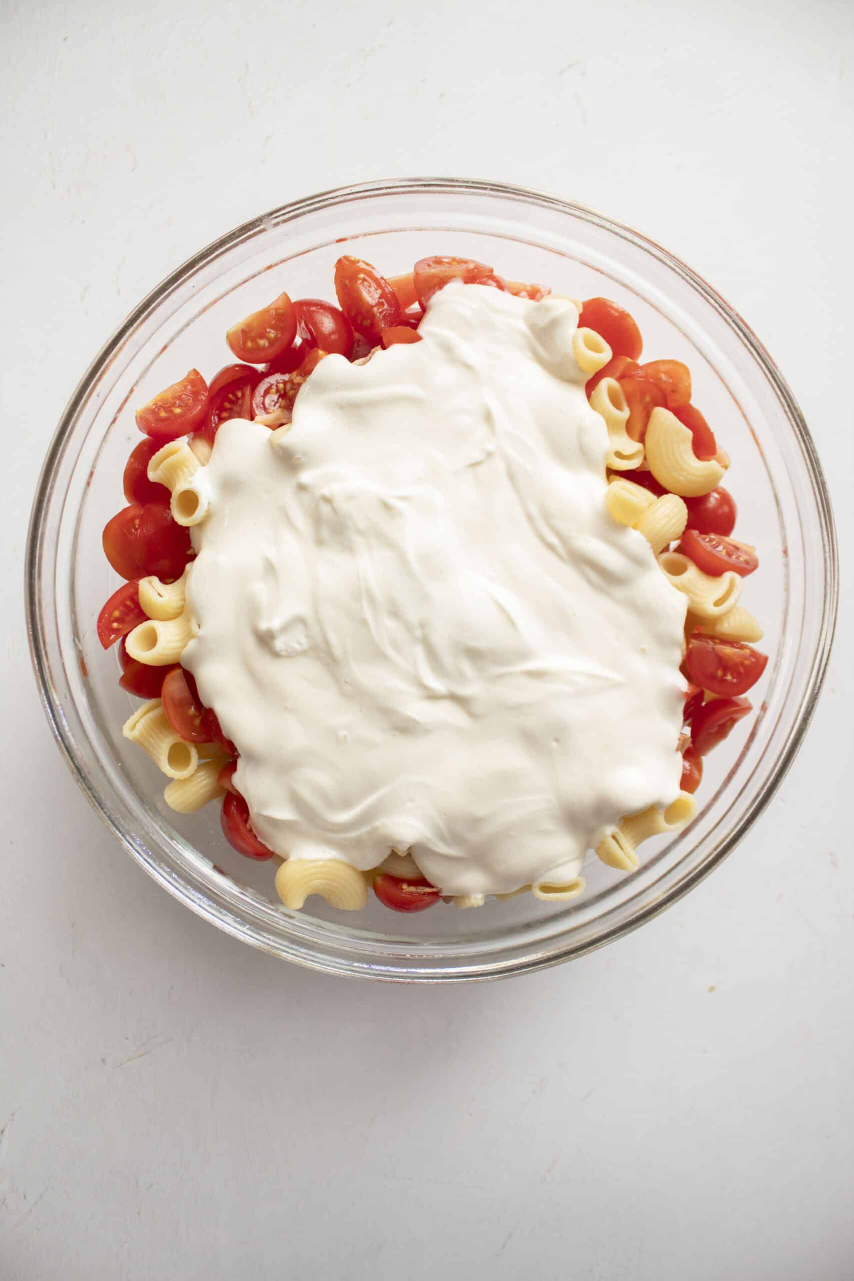 Cooked elbow macaroni in a large glass mixing bowl with quartered cherry tomatoes and diced bacon with a greek yogurt and mayo mixture.