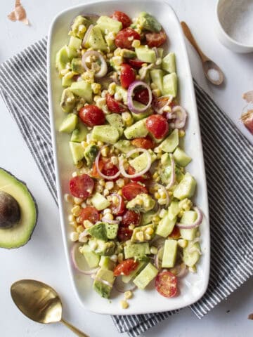 A platter of a corn and avocado salad with tomatoes, shallots, and cucumber with a napkin underneath and a gold spoon to the side.