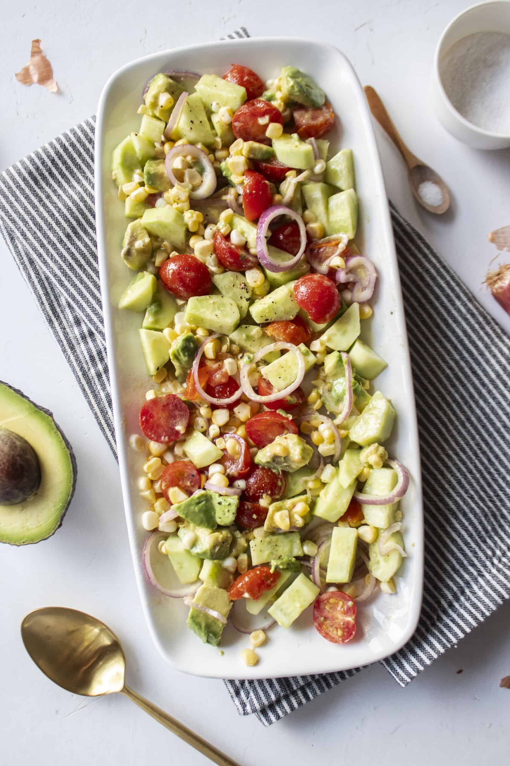 A platter of a corn and avocado salad with tomatoes, shallots, and cucumber with a napkin underneath and a gold spoon to the side.