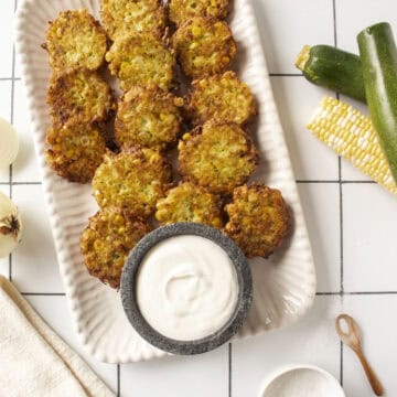 A platter of corn and zucchini fritters with a greek yogurt lime dip in a small marble bowl surrounded by zucchini and corn on the cob.