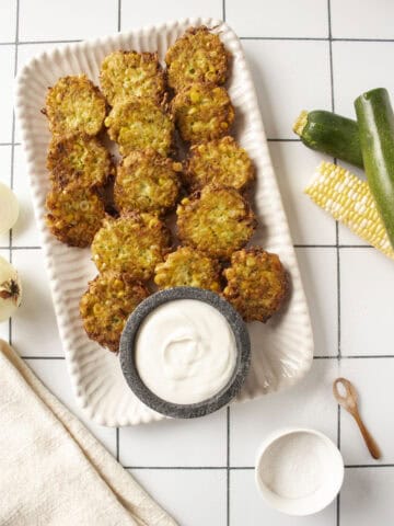 A platter of corn and zucchini fritters with a greek yogurt lime dip in a small marble bowl surrounded by zucchini and corn on the cob.