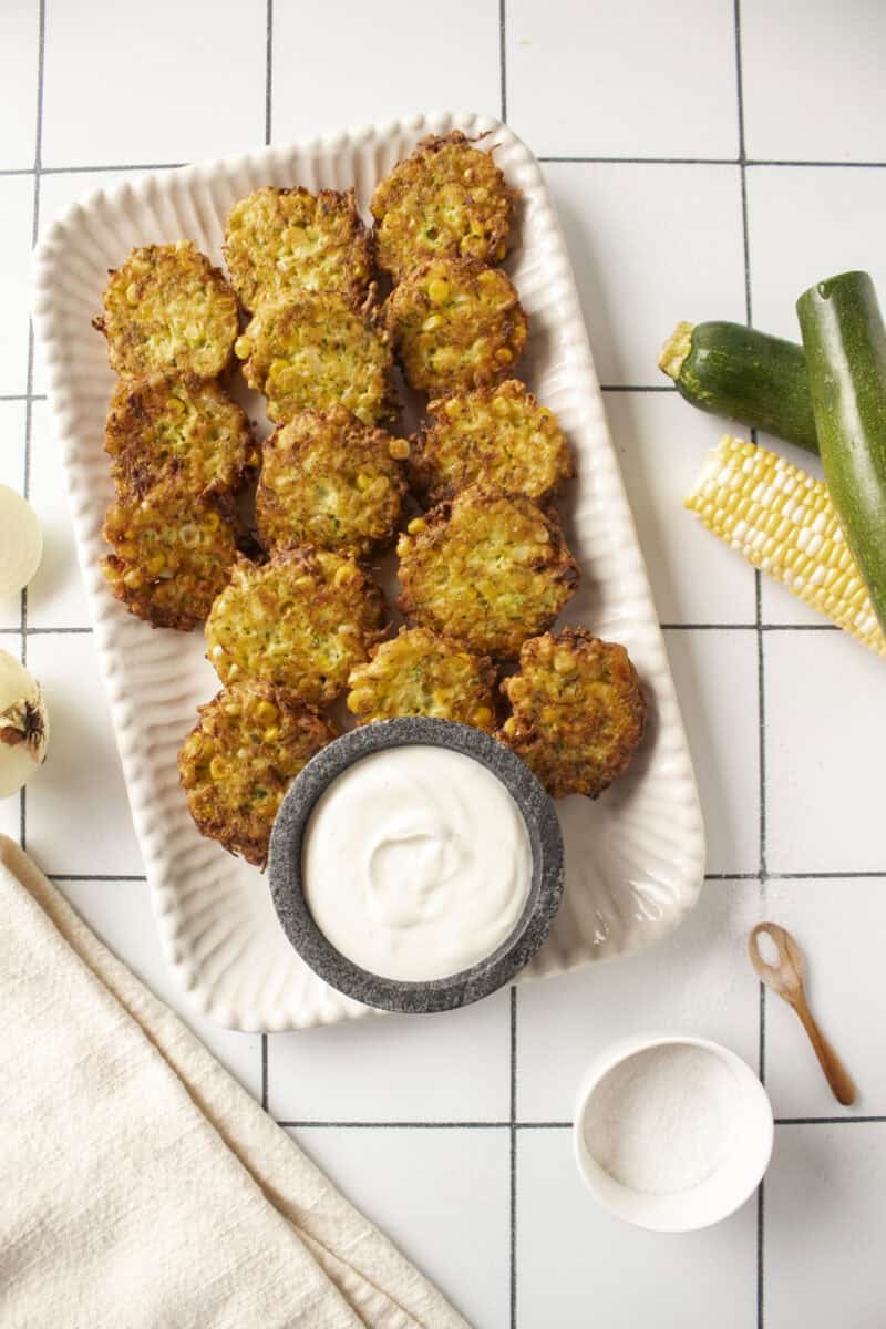 A platter of corn and zucchini fritters with a greek yogurt lime dip in a small marble bowl surrounded by zucchini and corn on the cob.