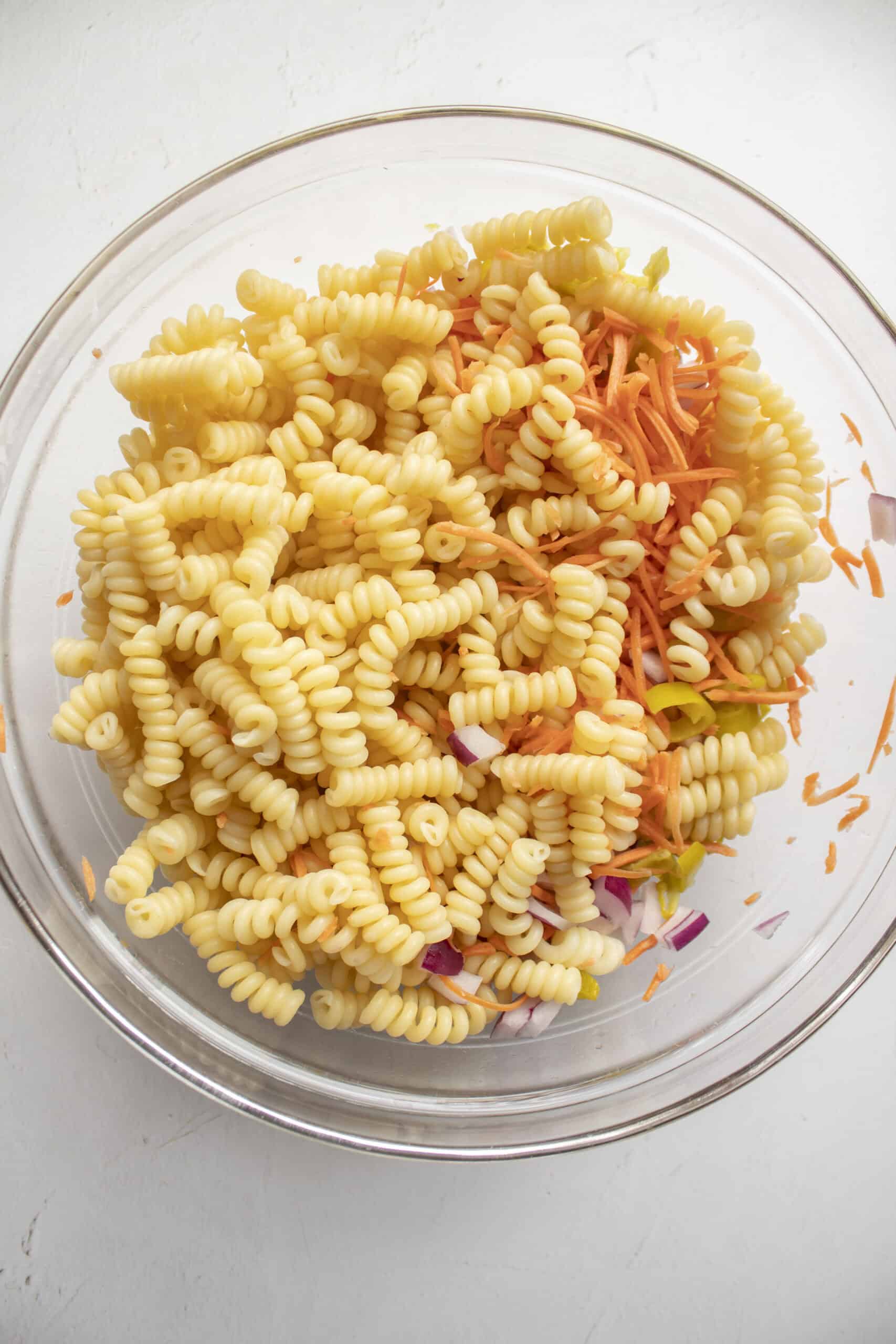 Cooked then cooled pasta in a large glass bowl mixed with carrots and pepperoncini.