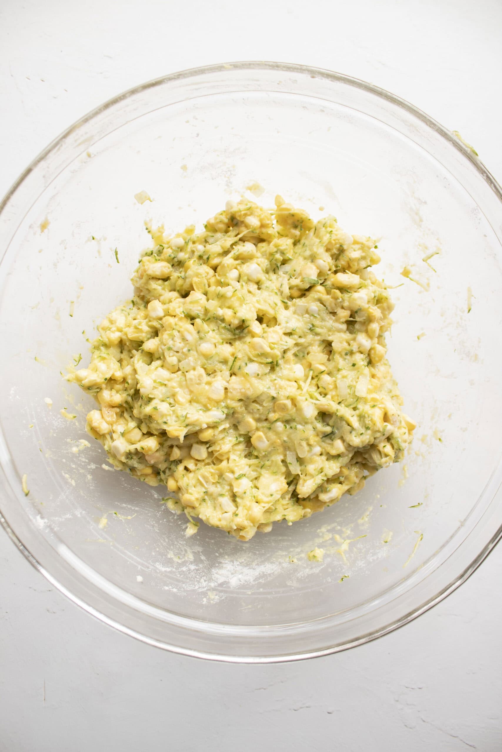 Corn, shredded zucchini, eggs, parmesan cheese, flour, garlic, powder, and diced onion in a large glass mixing bowl mixed together to form corn fritter batter.