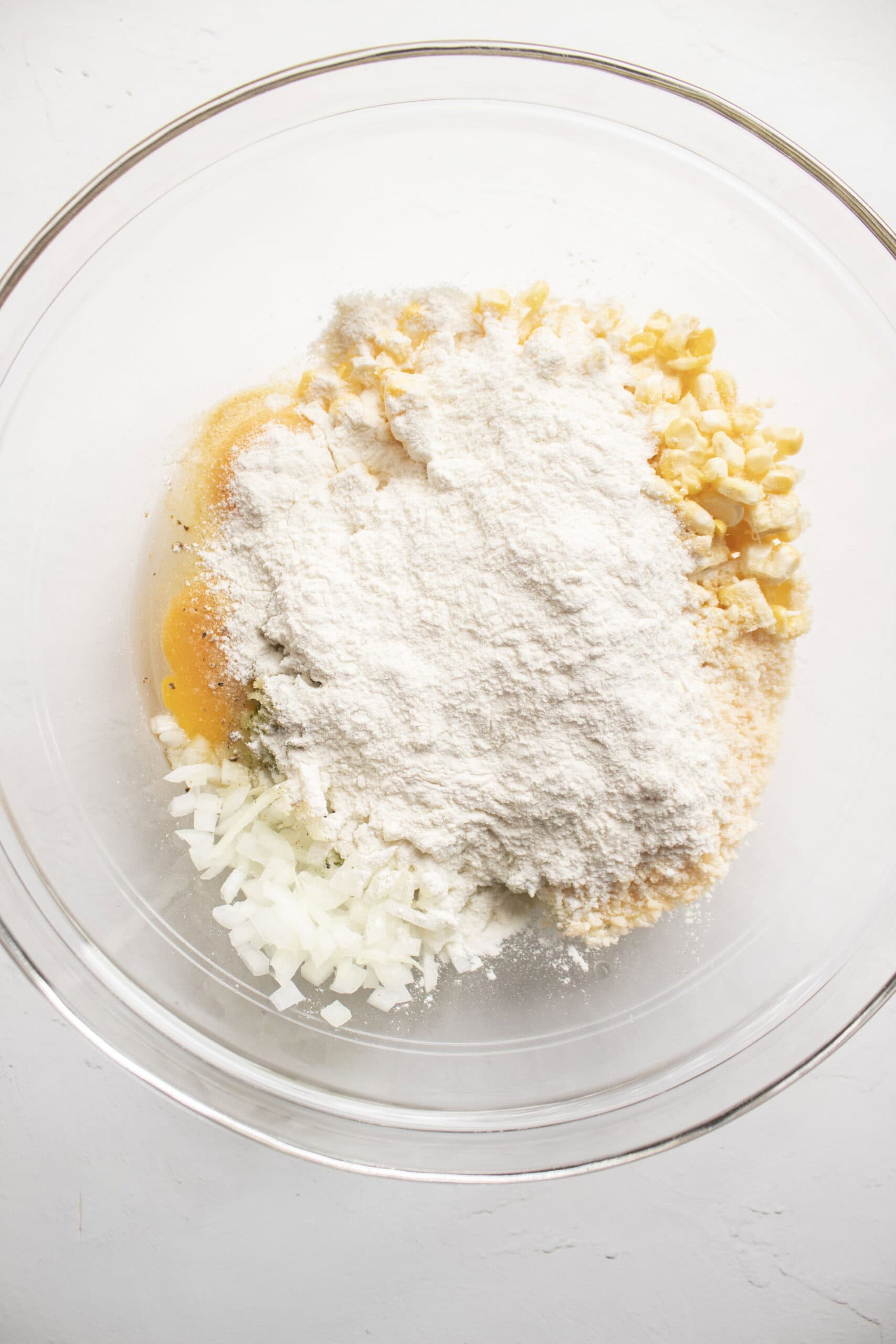 Corn, shredded zucchini, eggs, parmesan cheese, flour, garlic, powder, and diced onion in a large glass mixing bowl.