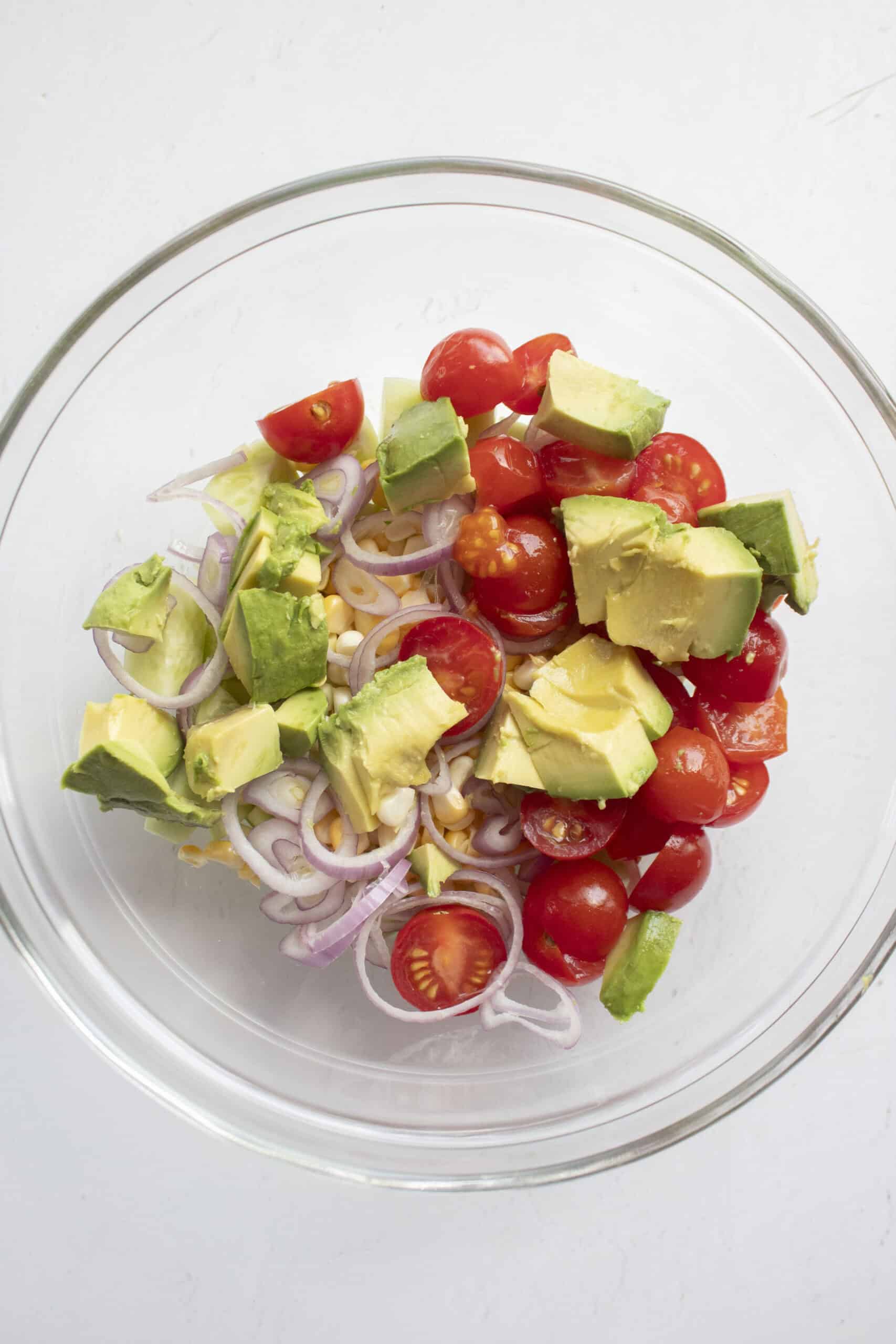 Halved grape tomatoes, corn, sliced shallots, and diced cucumbers in a glass mixing bowl topped with avocado.