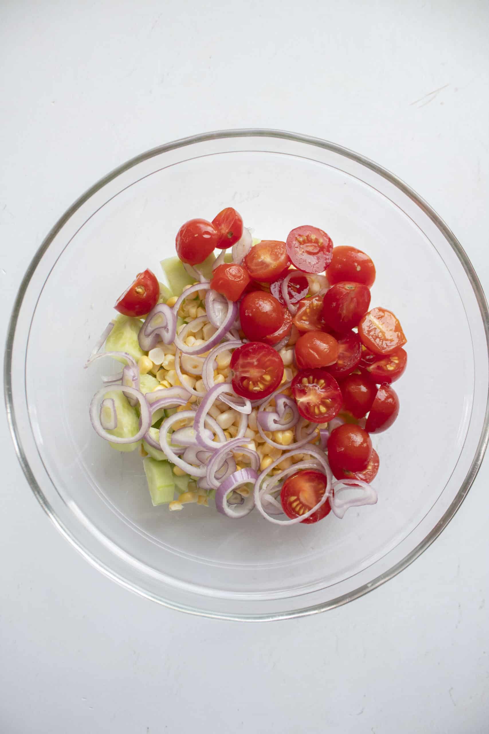 Halved grape tomatoes, corn, sliced shallots, and diced cucumbers in a glass mixing bowl.