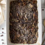 Baked babka bread pudding in a casserole dish surrounded by a napkin, serving spoons, and chocolate chips.