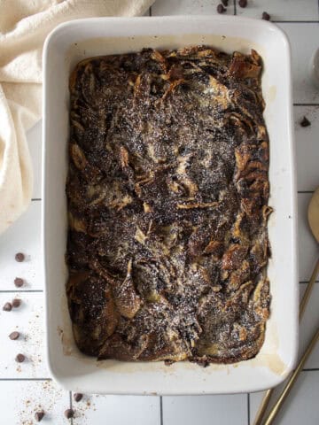 Baked babka bread pudding in a casserole dish surrounded by a napkin, serving spoons, and chocolate chips.