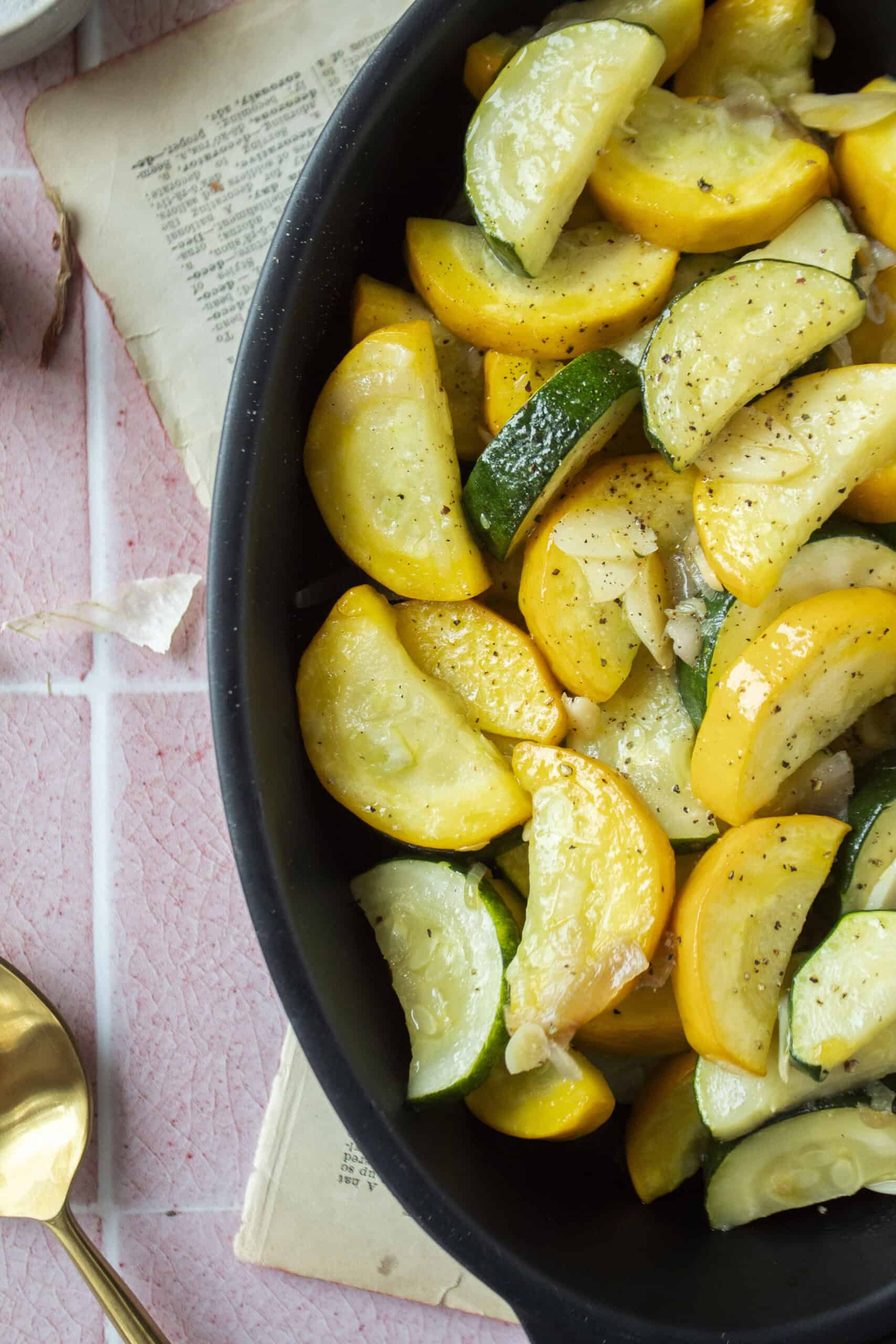 Close up of sauteed zucchini and yellow squash with shallots and garlic in a black enameled dish on a pink background.