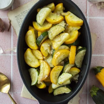Sauteed zucchini and yellow squash with shallots and garlic in a black enameled dish on a pink background.