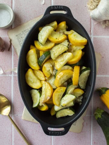 Sauteed zucchini and yellow squash with shallots and garlic in a black enameled dish on a pink background.