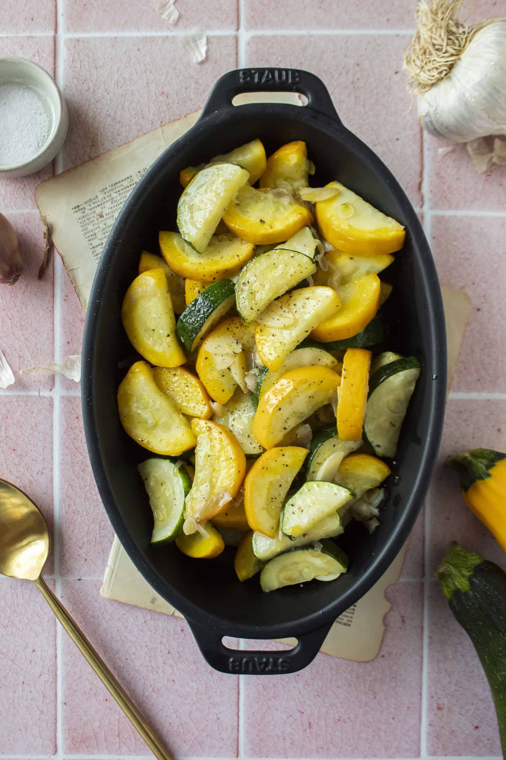 Sauteed zucchini and yellow squash with shallots and garlic in a black enameled dish on a pink background.