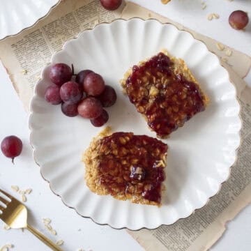 Two slices of peanut butter oatmeal bake topped with grape jelly on a scalloped plate.