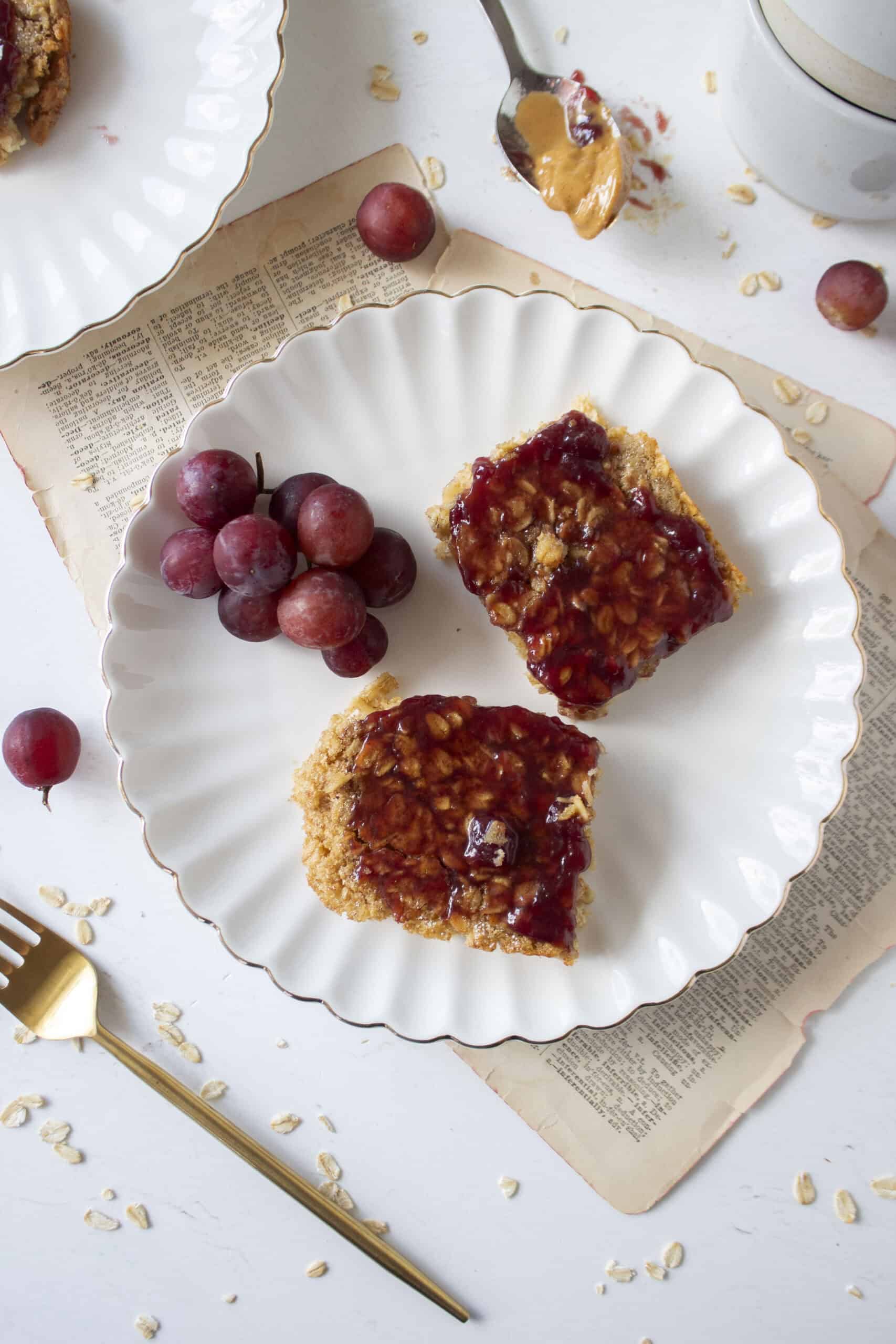 Two slices of peanut butter oatmeal bake topped with grape jelly on a scalloped plate.