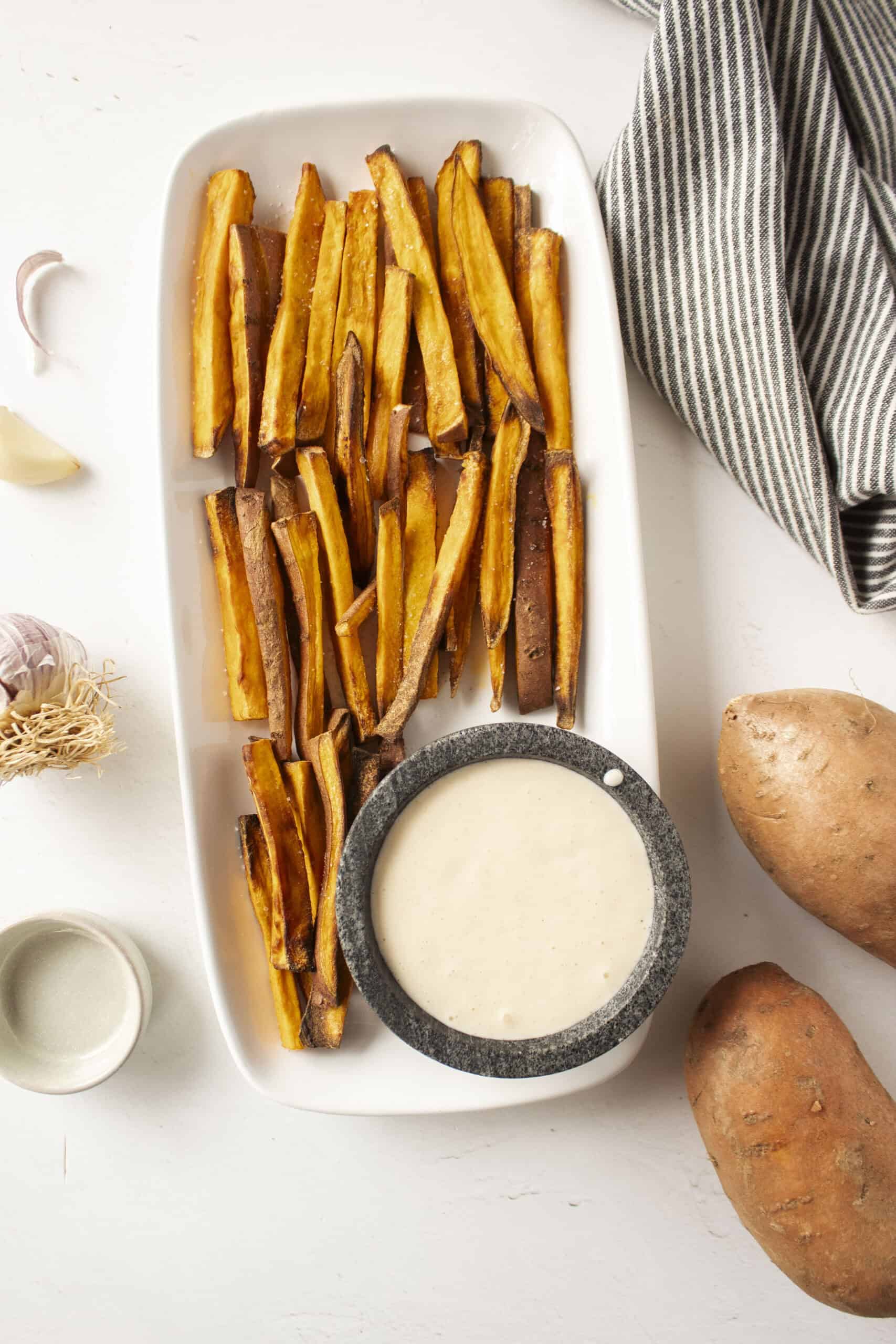 A white plate of air fryer sweet potato fries with a small bowl of garlic aioli surrounded by a blue and white striped napkin and sweet potatoes.