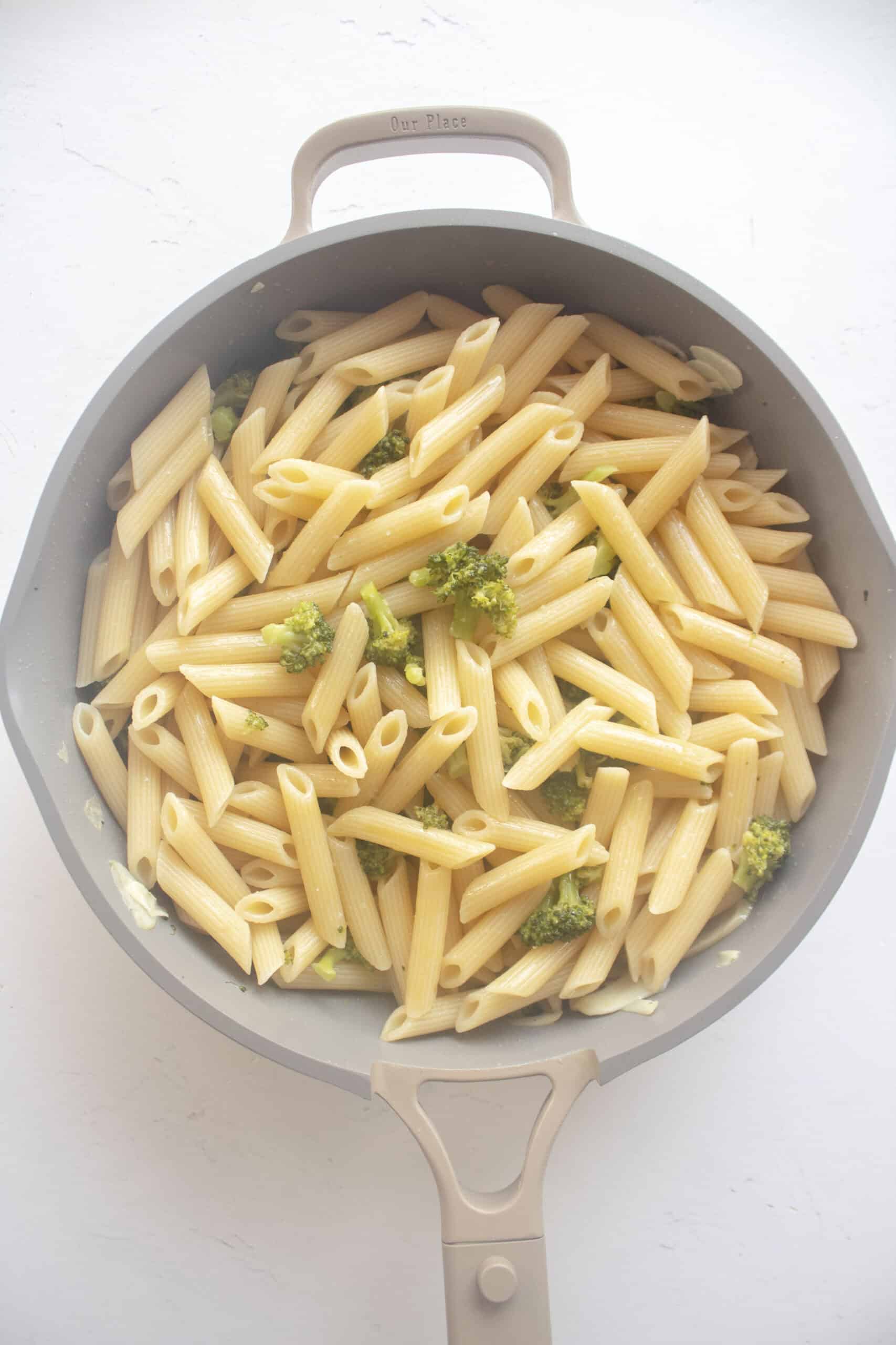 Cooked penne pasta mixed with blanched broccoli tossed with garlic in a frying pan with olive oil.