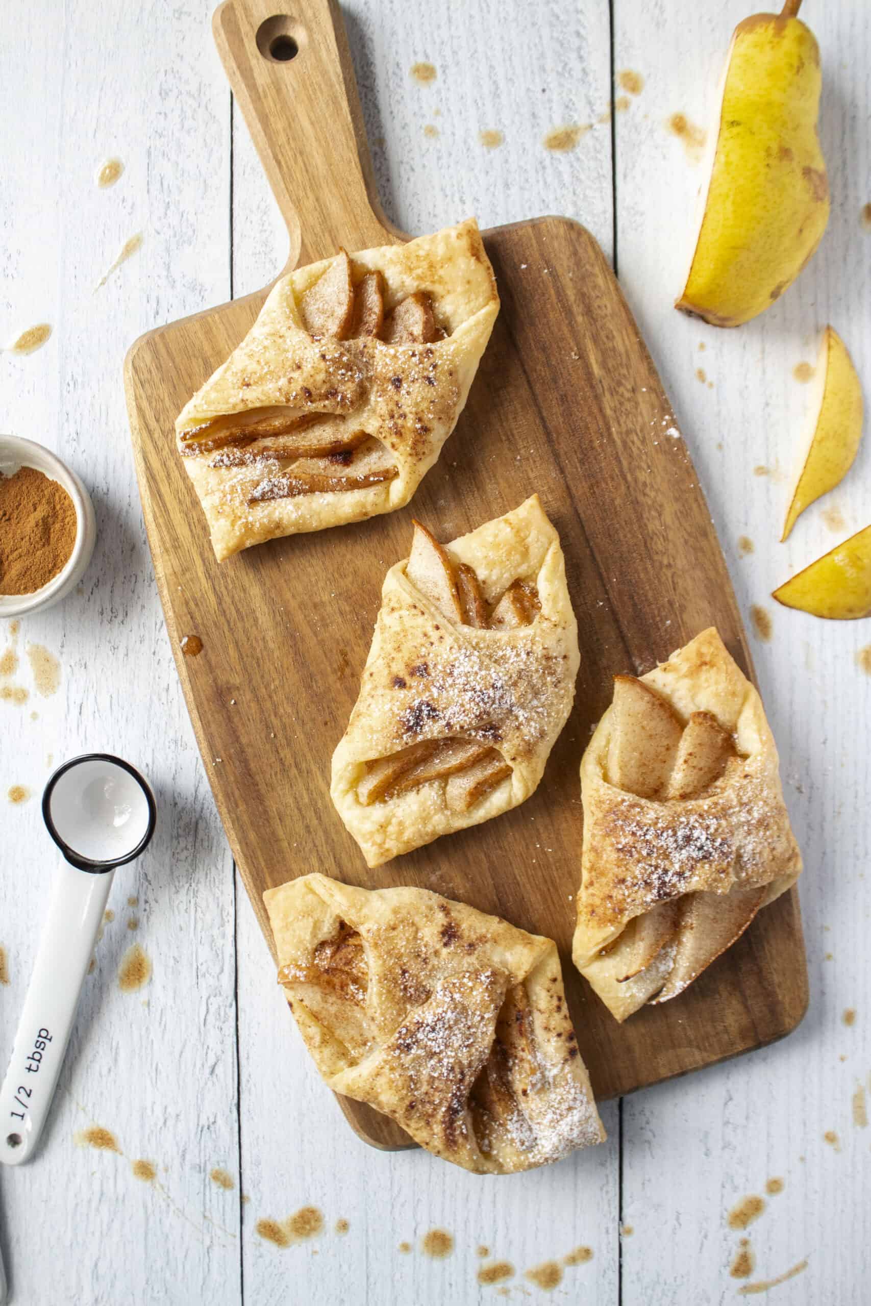Four pear and brown butter turnovers on a wooden serving board surrounded by sliced pear and measuring spoons.