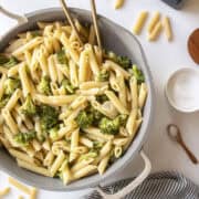 Penne pasta with garlic broccoli in a frying pan with two golden spoons in the pan surrounded by a napkin, salt, and dried pasta.