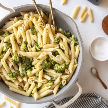 Penne pasta with garlic broccoli in a frying pan with two golden spoons in the pan surrounded by a napkin, salt, and dried pasta.